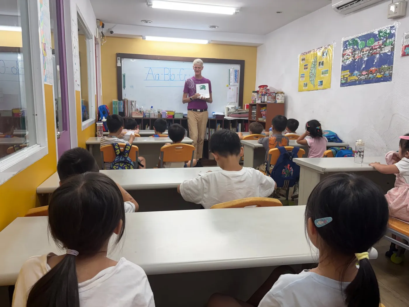 老師在寫有 A、B、C 的白板前展示青蛙圖卡，學生背對鏡頭上課